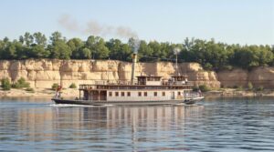Vintage gas-powered riverboat from early 1900s cruising on a calm river with tree-lined sandstone bluffs in the background under soft daylight
