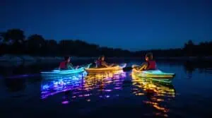 Three friends in LED-lit clear kayaks paddle on a calm river at night, their boats casting colorful reflections on the water with dim moonlight, distant trees, and rocks in the background.