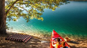 Two people kayak toward a secluded lakeside cove with a picnic blanket and basket under pine trees on a calm, sunlit lake.