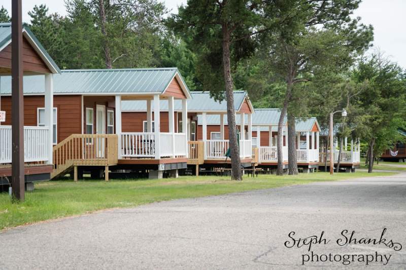 A row of small wooden cabins with porches lines a paved path, surrounded by grass and trees.