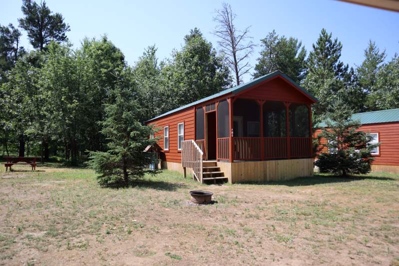 A small red cabin with a screened porch sits on a grassy lot surrounded by trees, with a fire pit and a picnic table nearby.