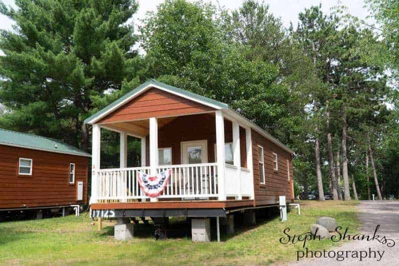 A small brown cabin with a covered front porch and white railings, decorated with a red, white, and blue banner, set among trees in a wooded area.