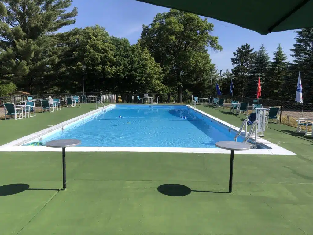 Rectangular outdoor swimming pool with clear water, surrounded by green deck, tables, chairs, and trees in the background under a sunny sky.