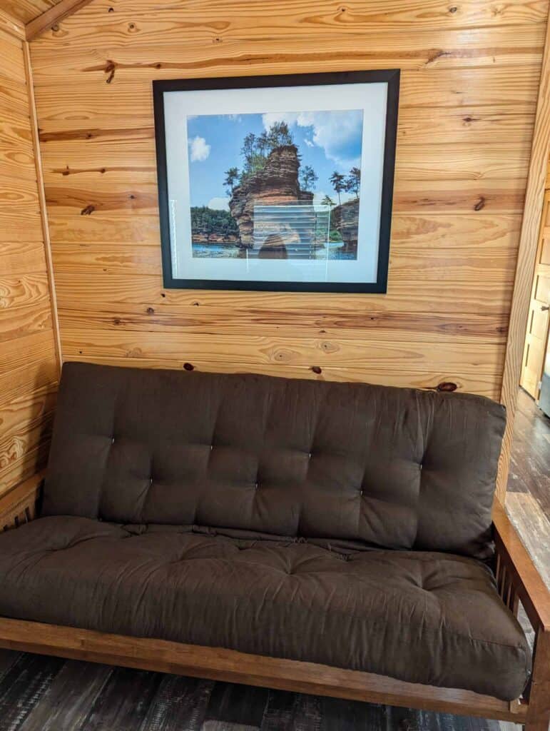 A brown futon sits against a wooden wall with a framed photograph of a rock formation over water hanging above it.