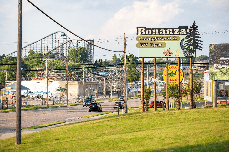 A road with cars passes by a sign for Bonanza Campground & RV Park; a large roller coaster is visible in the background.