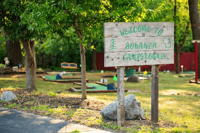 A wooden sign reading "Welcome to Bonanza Campground" stands in front of a mini golf course surrounded by trees and grass.