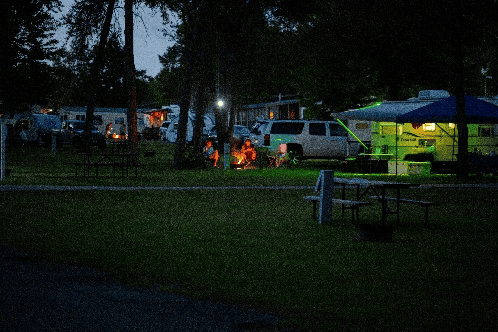 People are sitting around a campfire at dusk in a campground with tents, parked vehicles, and picnic tables visible in the background.