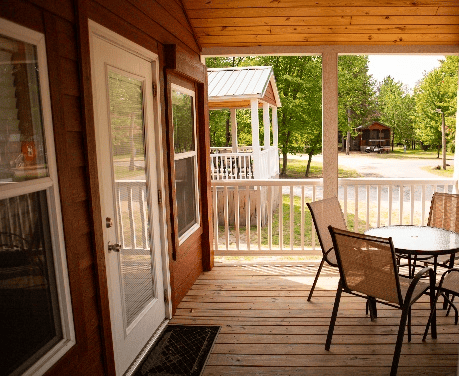 A wooden porch with a glass door, table, and four chairs overlooks a gravel path, trees, and a small gazebo in the background on a sunny day.