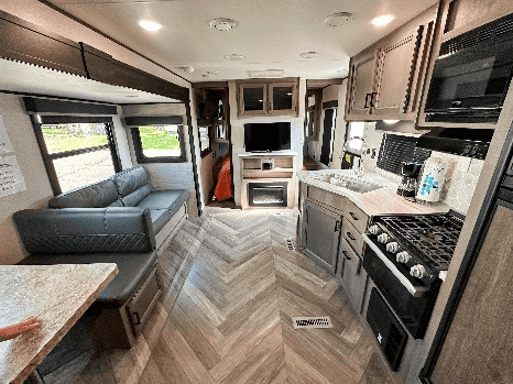 Interior view of a modern RV with a sofa, dining area, kitchen, television, and fireplace, featuring wood-look flooring and neutral-toned decor.