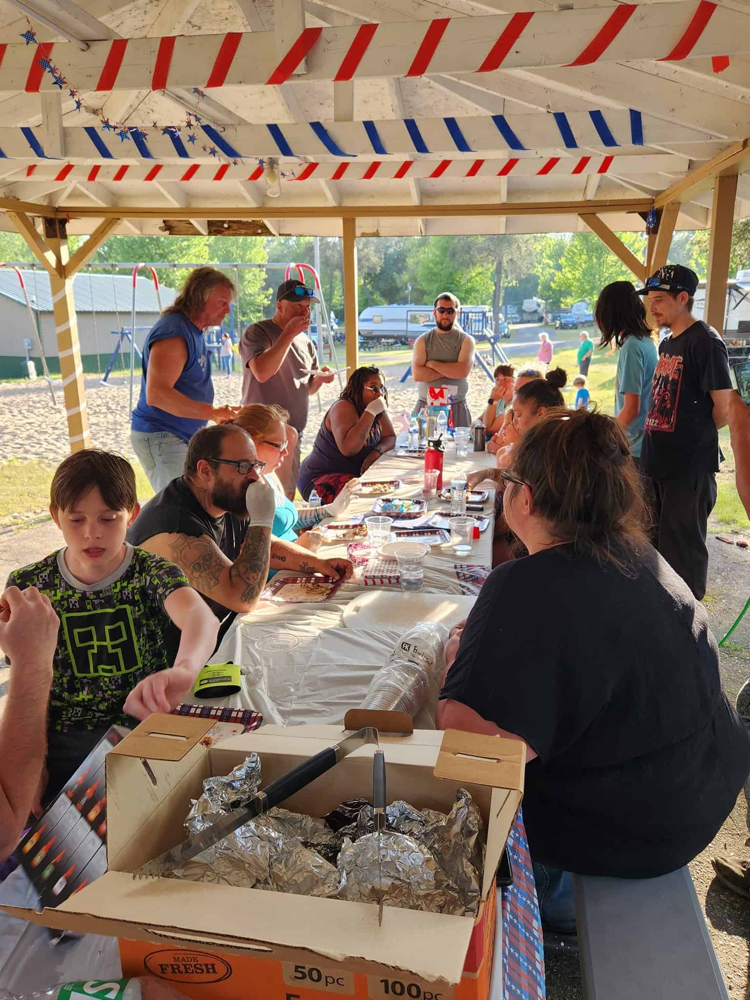 A group of people sit and stand around a long picnic table covered with food and supplies under a pavilion at a park on a sunny day.