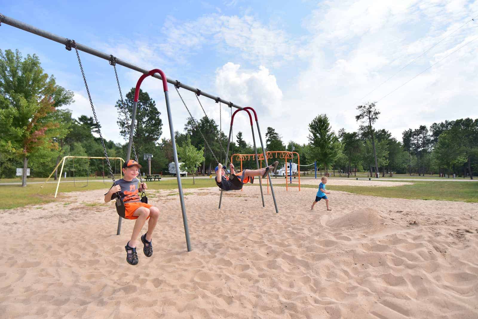 Three children play on a sandy playground; two are on swings while one runs nearby. Trees and more playground equipment are in the background.