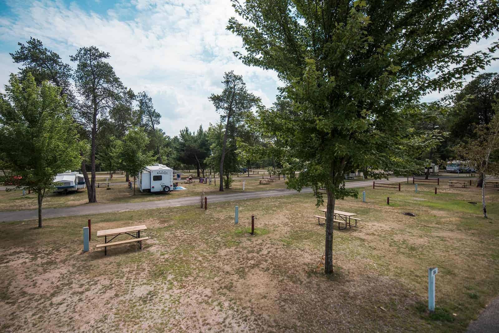 Campground with scattered picnic tables, a few RVs, and tall trees under a partly cloudy sky. Some empty camping spots are visible.