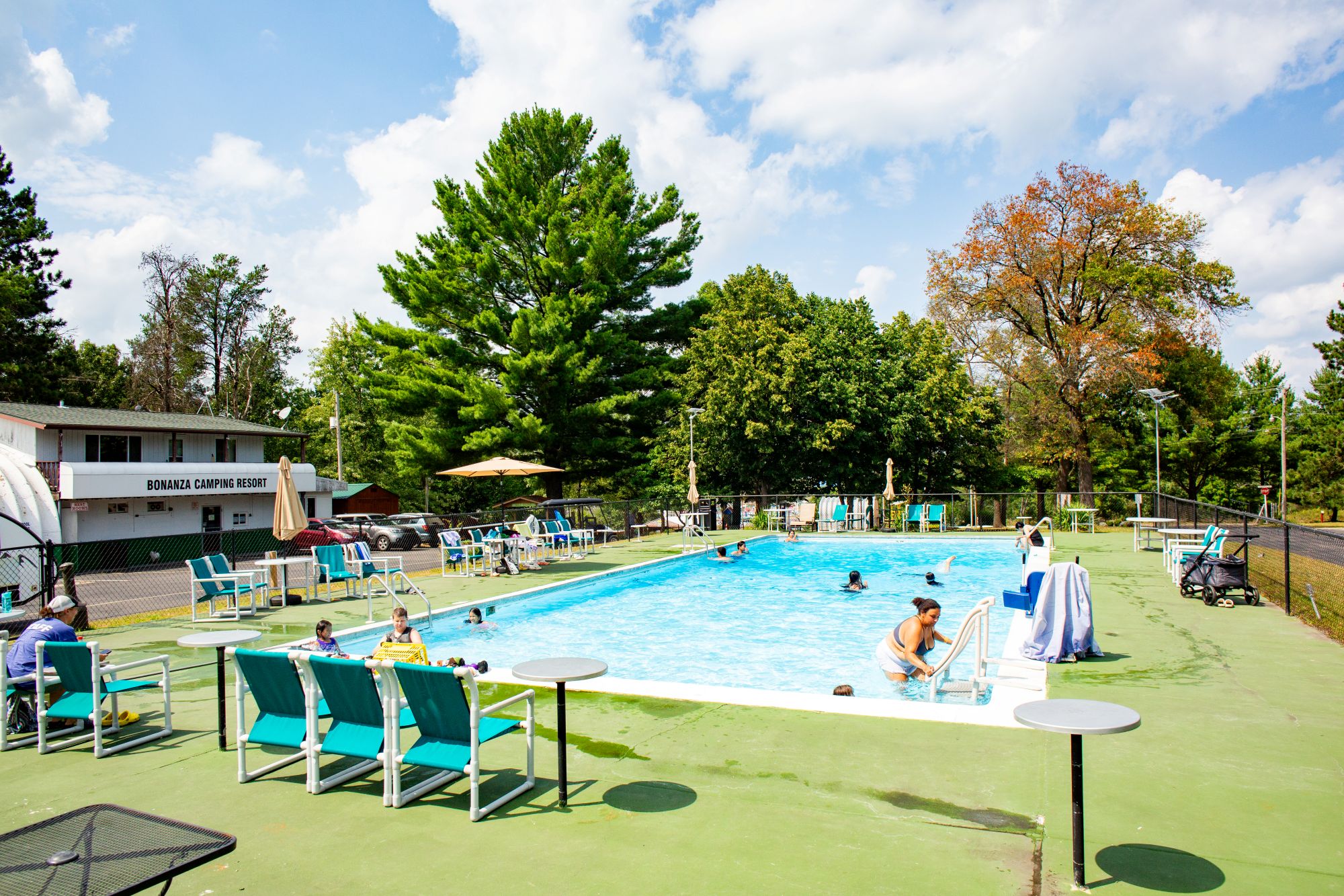 Outdoor swimming pool at a campsite with people swimming and lounging, surrounded by trees and a building labeled "Bonanza Camping Resort.