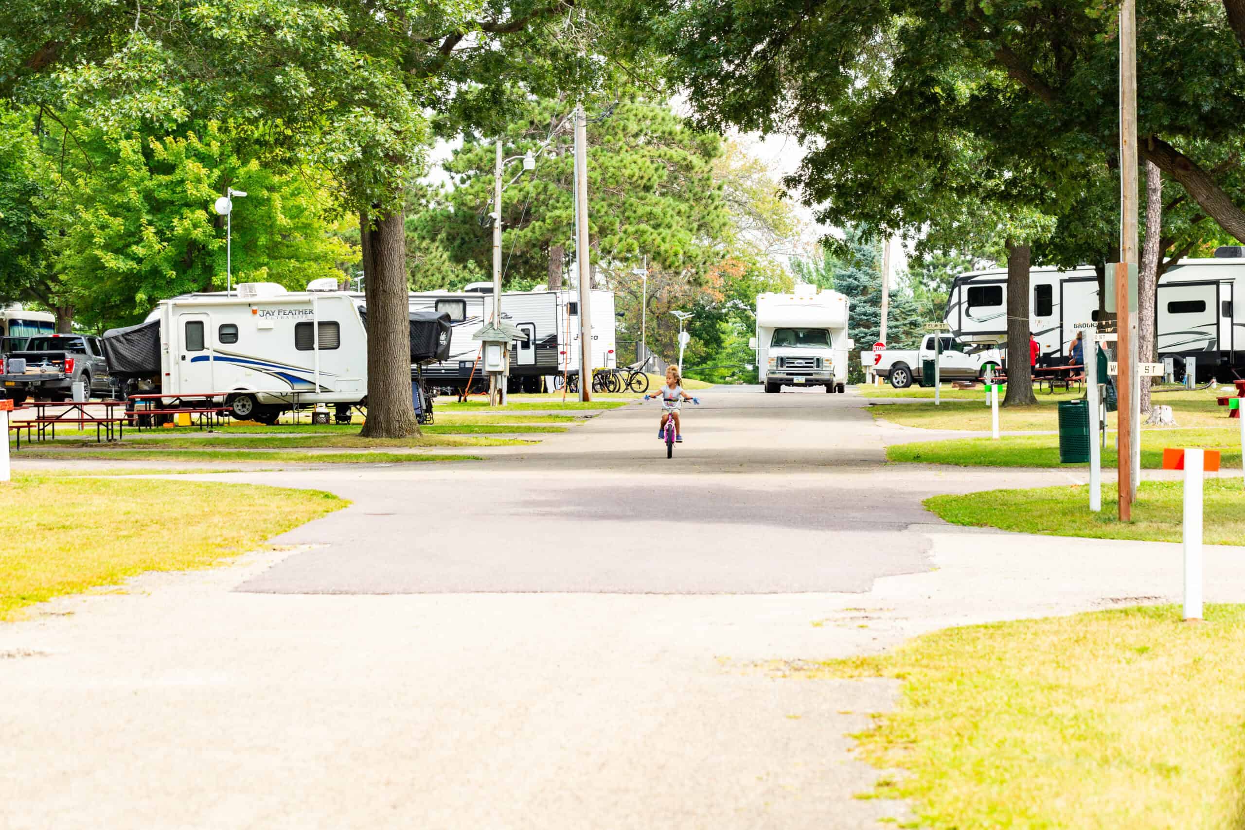 A young child rides a bike on a paved path through a campground with parked RVs and trees in the background.