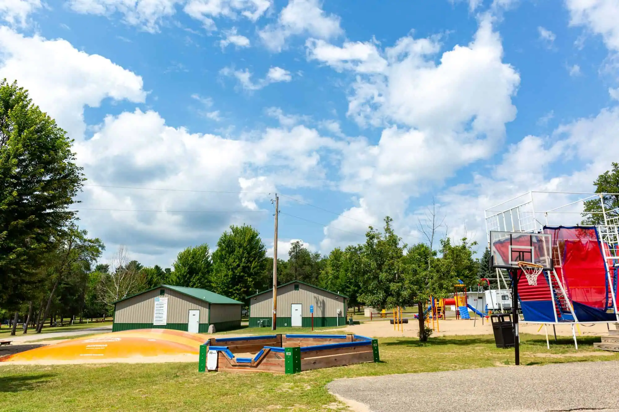 Outdoor recreation area with a basketball hoop, large inflatable play mound, lawn games, playground, and green-roofed buildings surrounded by trees under a partly cloudy sky.