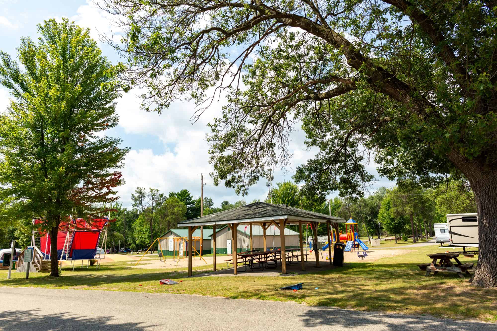 Outdoor picnic shelter with tables under a large tree, near a playground and red inflatable structure on a sunny day.