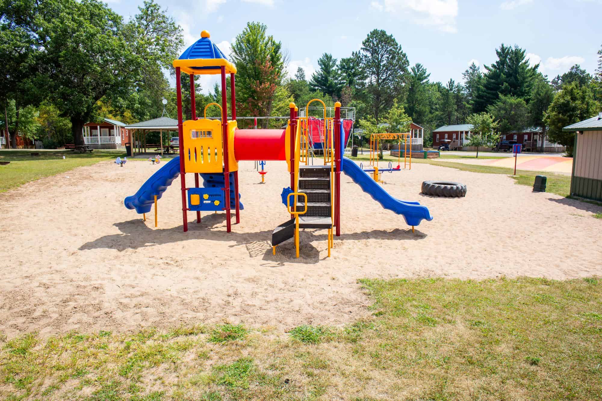 A playground with a slide, climbing steps, and a tunnel on sandy ground, surrounded by grass, trees, and nearby buildings under a partly cloudy sky.