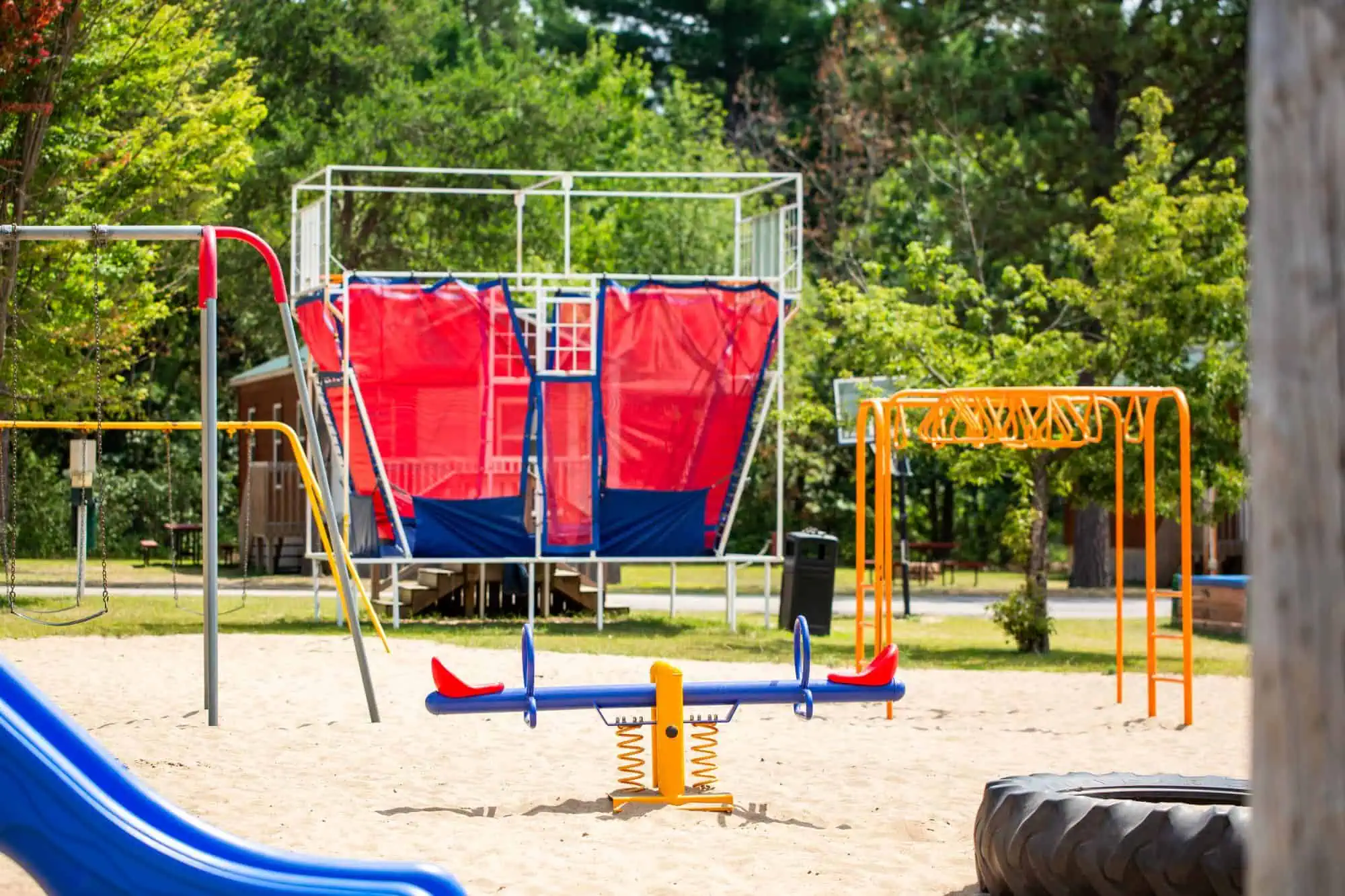 Playground with a seesaw, climbing structure, monkey bars, and a large play structure with red netting, surrounded by trees and sand.