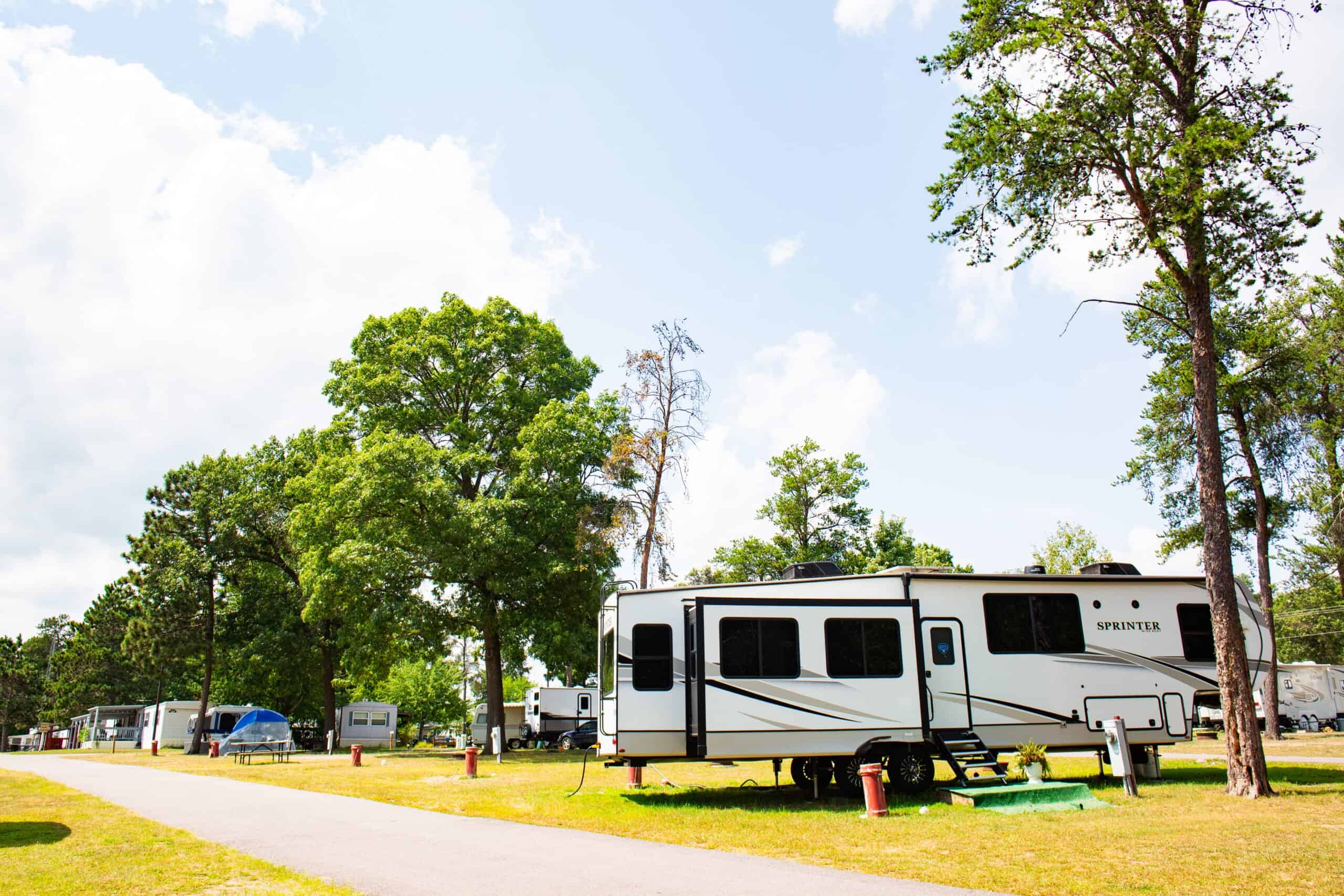 A white RV is parked on a grassy campsite near trees in a sunny RV park, with more RVs and a paved road visible in the background.