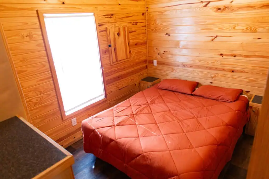 A small bedroom with wood-paneled walls, a window, a double bed with an orange comforter, two pillows, and built-in nightstands on each side.