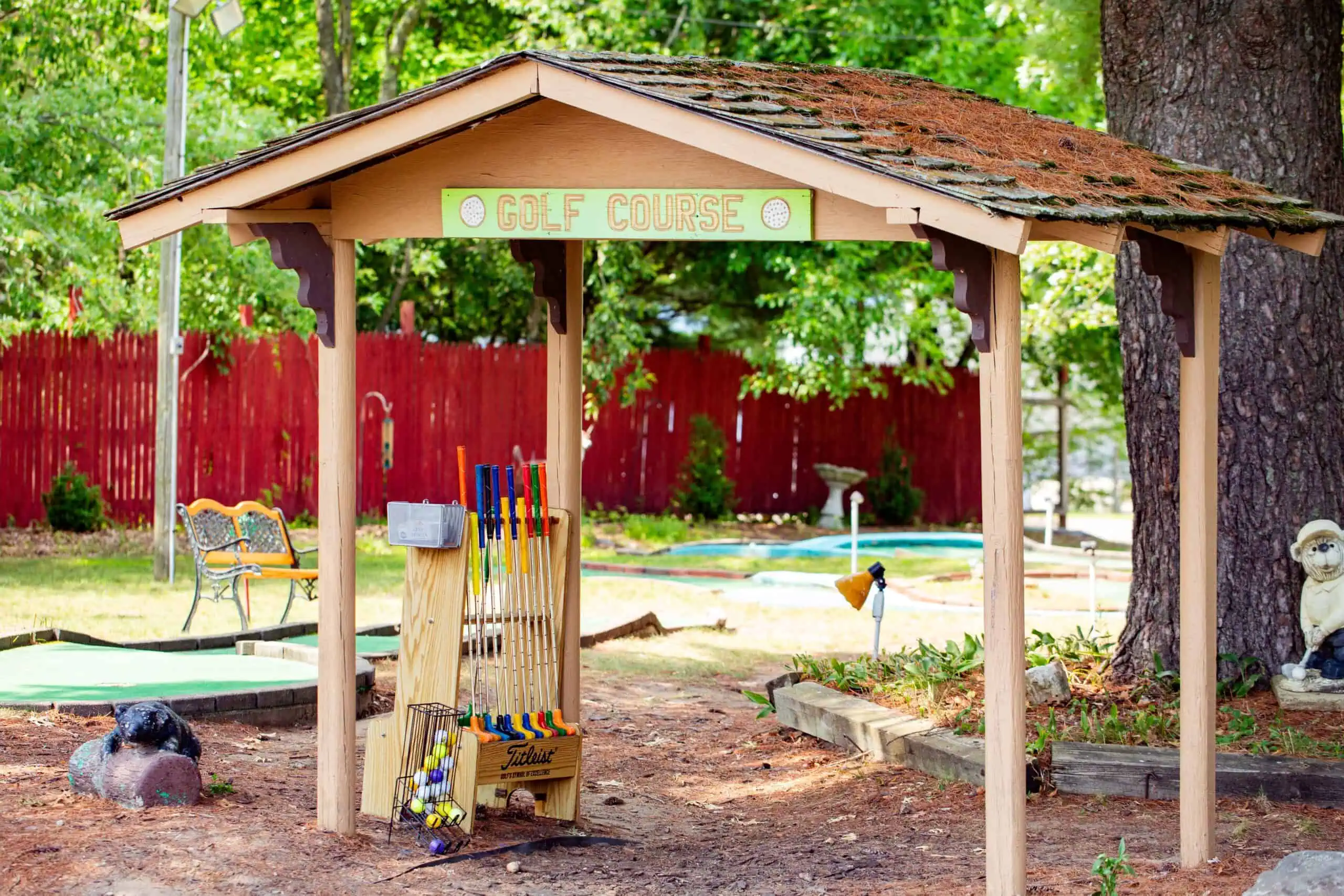 Wooden shelter labeled "Golf Course" with golf clubs and putters beneath it, set in a shaded outdoor mini-golf area with benches and trees in the background.