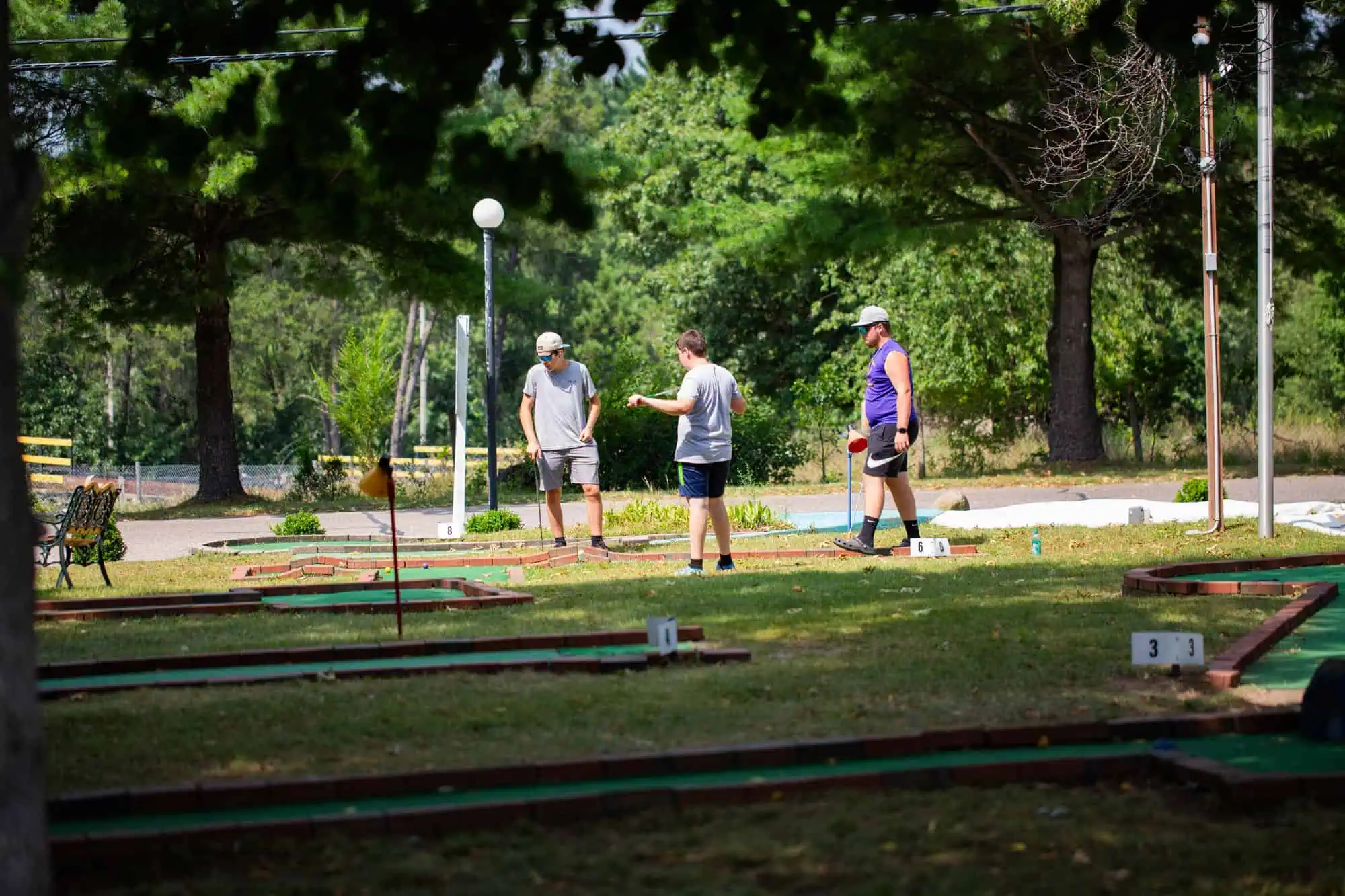 Three people stand on a mini golf course surrounded by trees and greenery, each holding a putter and appearing to discuss their next move.