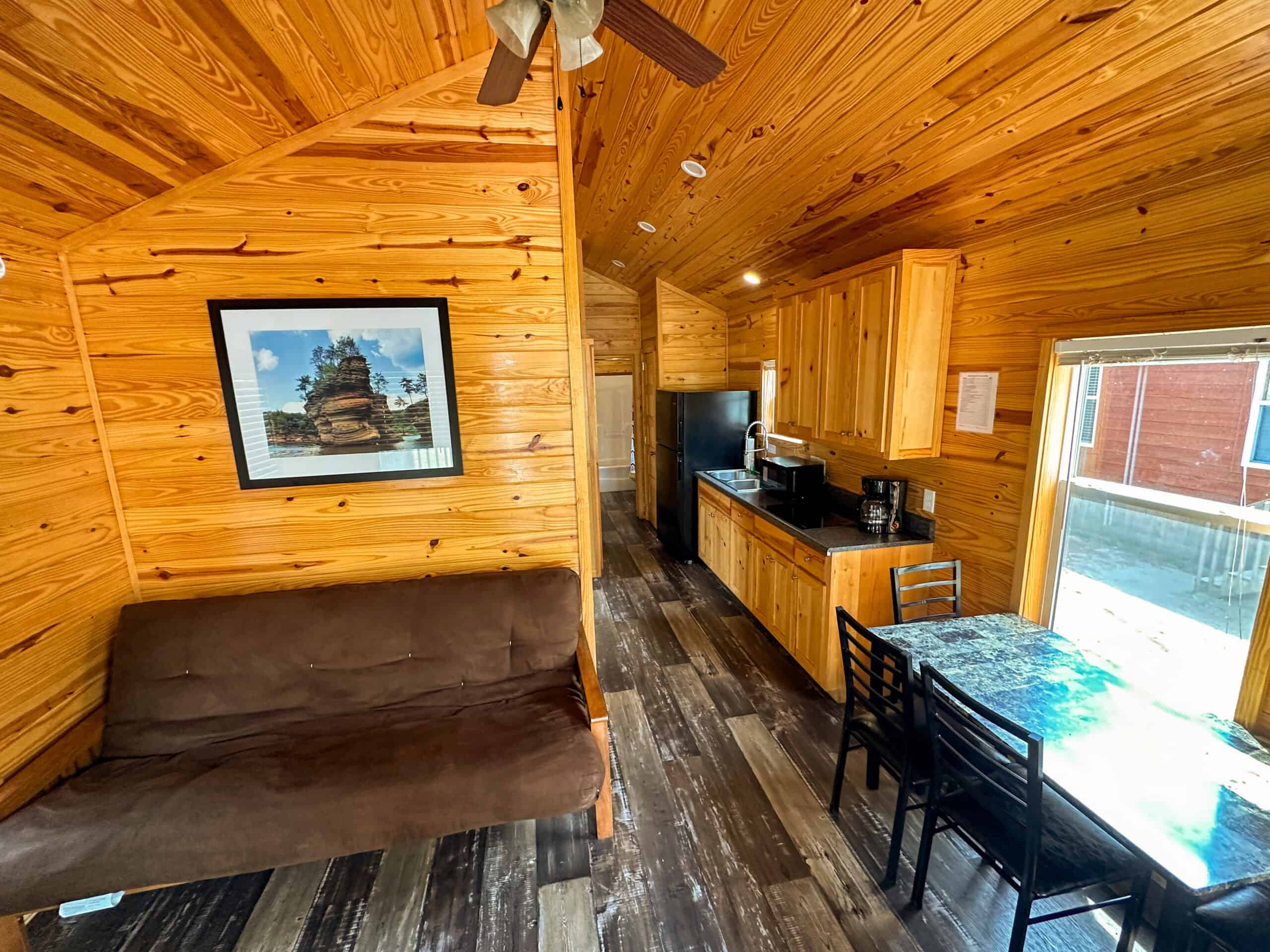 Small cabin interior with wooden walls and ceiling, a brown futon, framed photo, kitchen area with appliances, and a small dining table with chairs near a window.