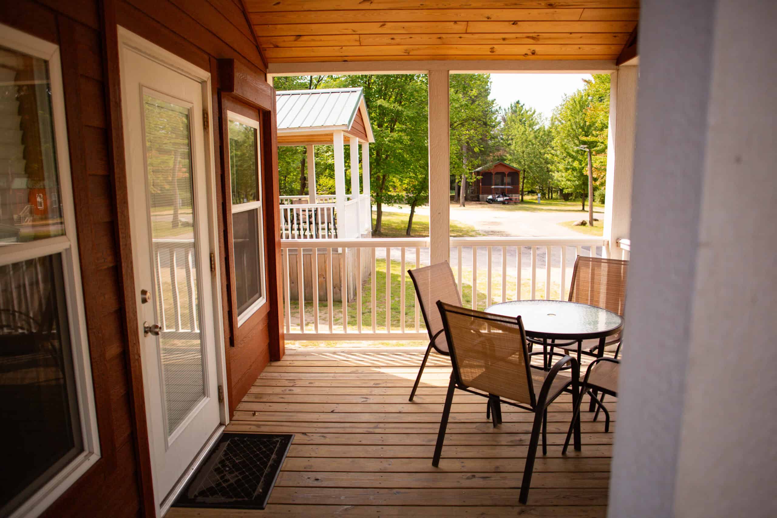 A wooden porch with a glass table and four chairs, white railing, and a view of a gazebo and cabins surrounded by trees.