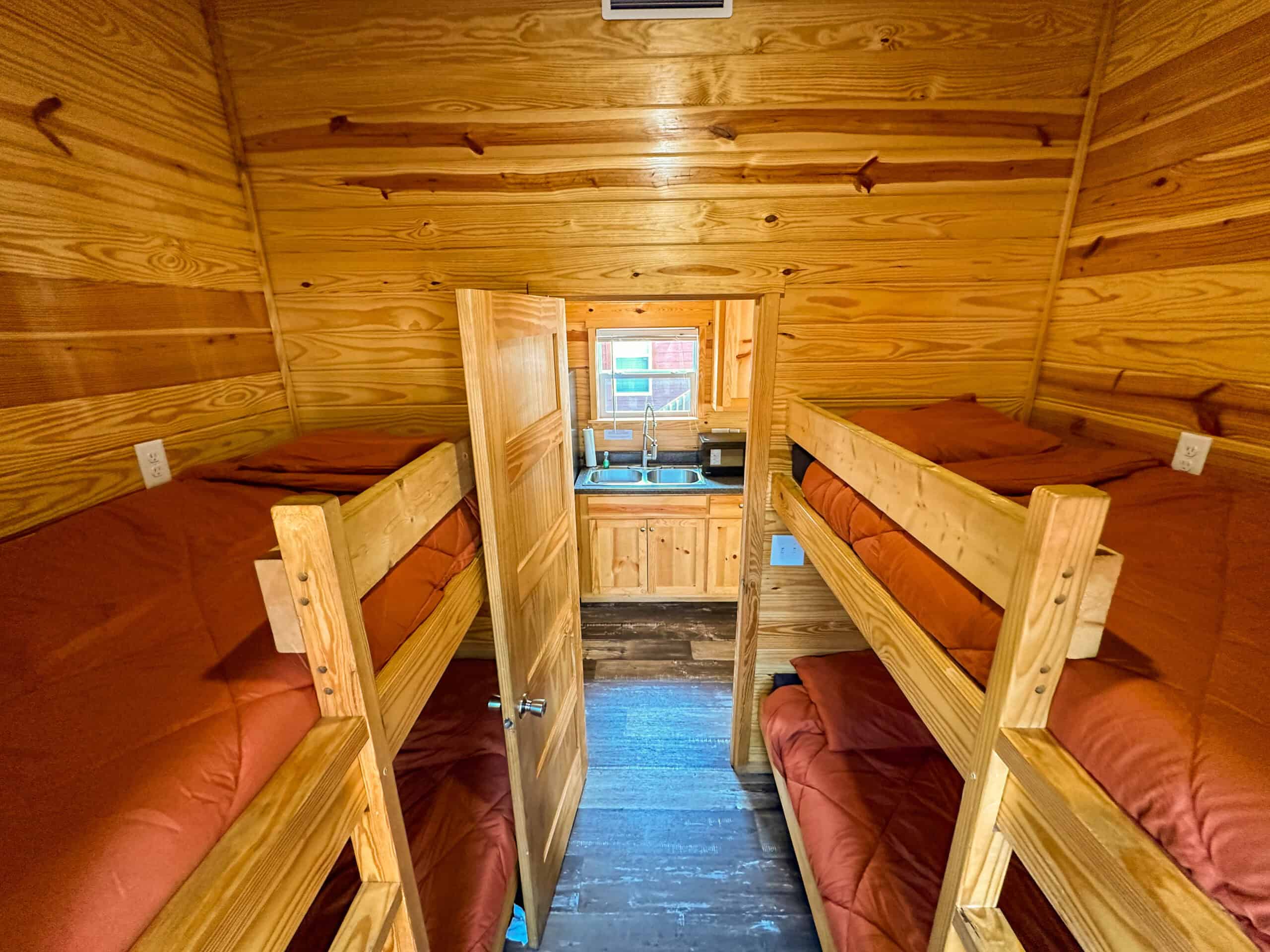 A small wooden cabin room with two sets of bunk beds and red bedding, viewed from the doorway, with a kitchenette visible in the background.