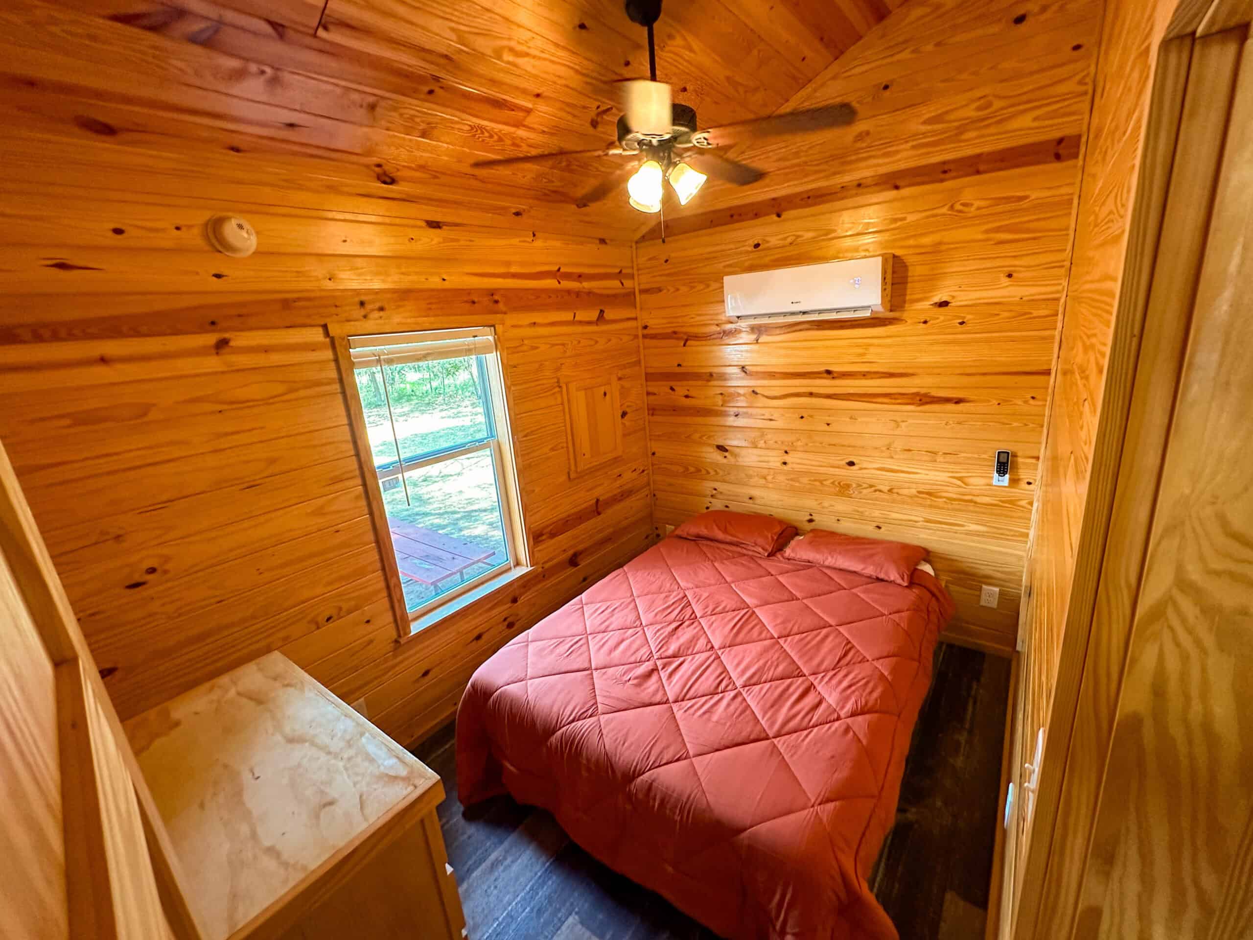 Small wooden bedroom with a bed covered in a red comforter, ceiling fan, wall-mounted air conditioner, dresser, and a window letting in natural light.