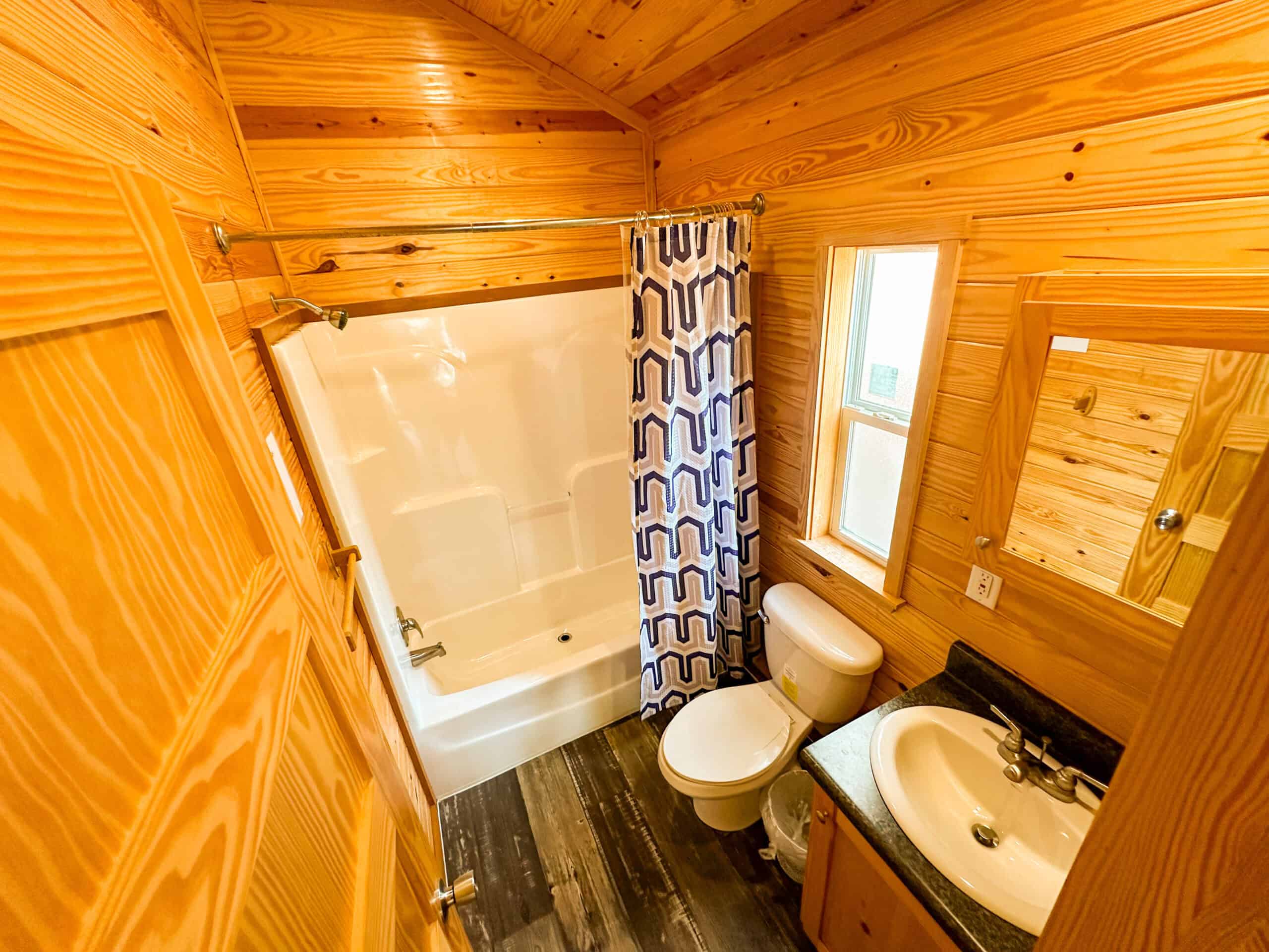 A small bathroom with wood-paneled walls, a bathtub with a geometric-patterned shower curtain, a toilet, a sink with a mirror, and a window.