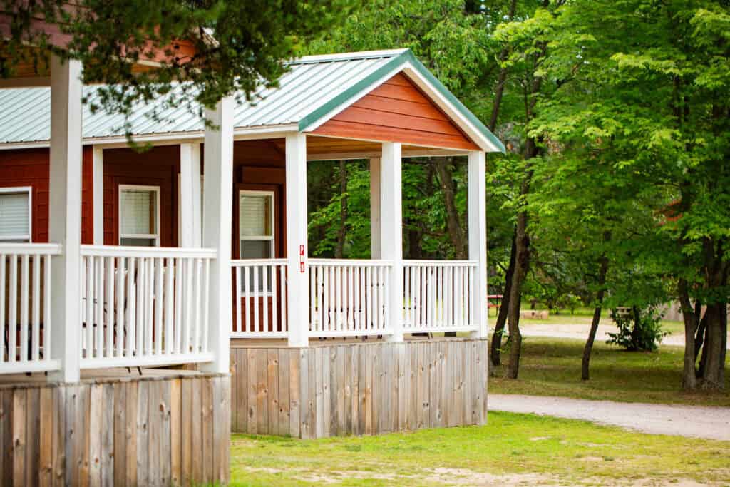 A small red cabin with a white porch and railing is surrounded by green trees and grassy ground.