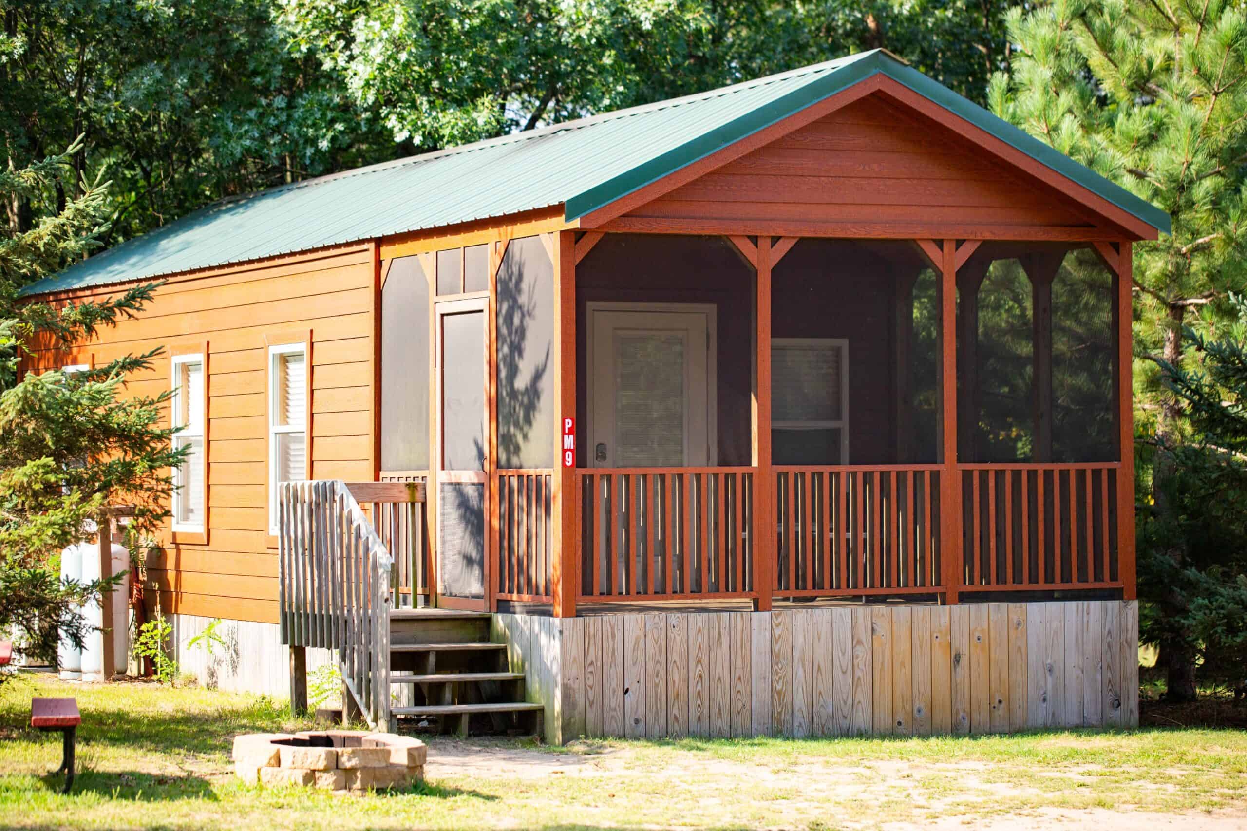 A small wooden cabin with a green metal roof and a screened front porch, surrounded by trees and grass, with steps leading up to the entrance.