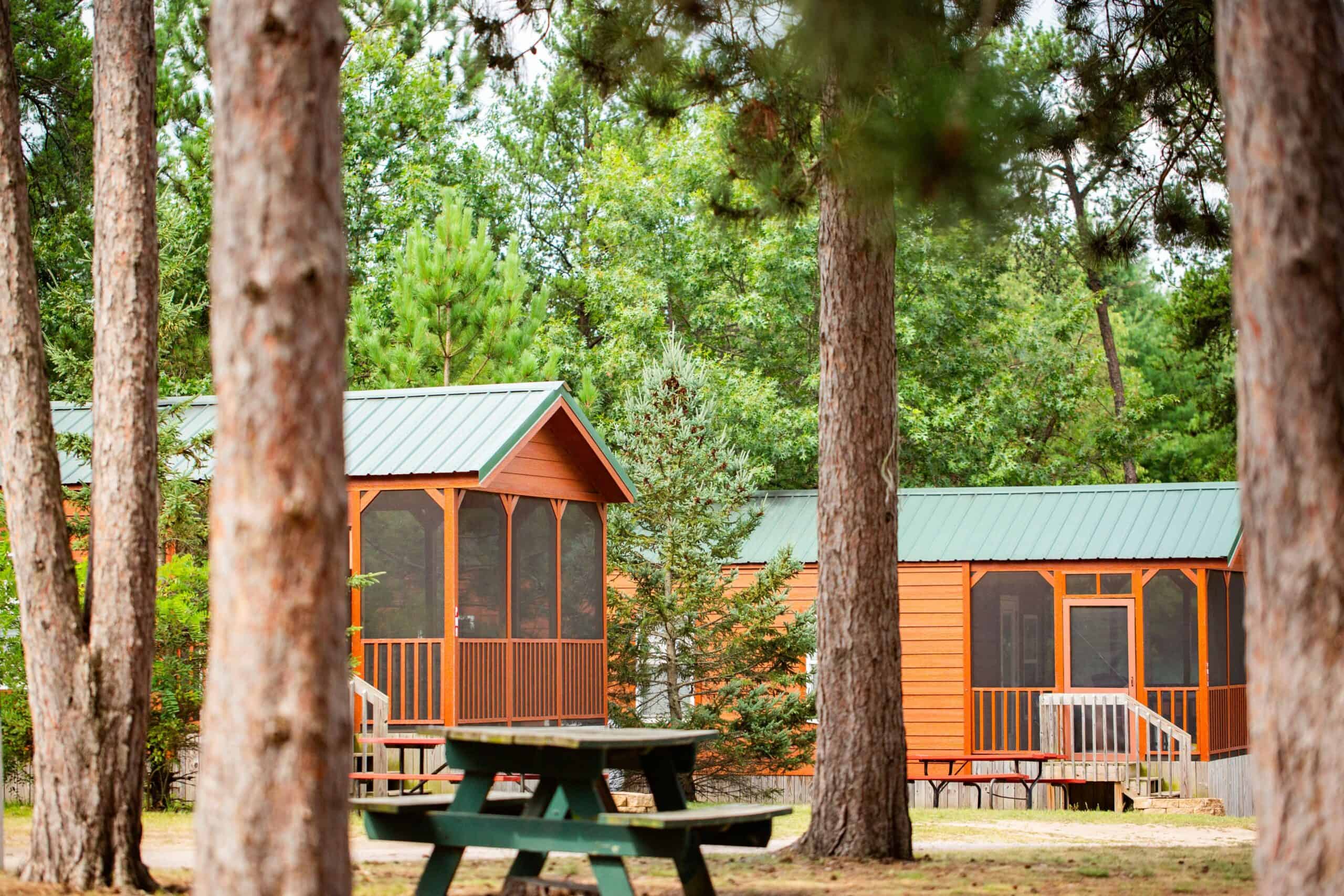 Wooden cabins with green metal roofs are surrounded by trees. A green picnic table sits in the foreground.