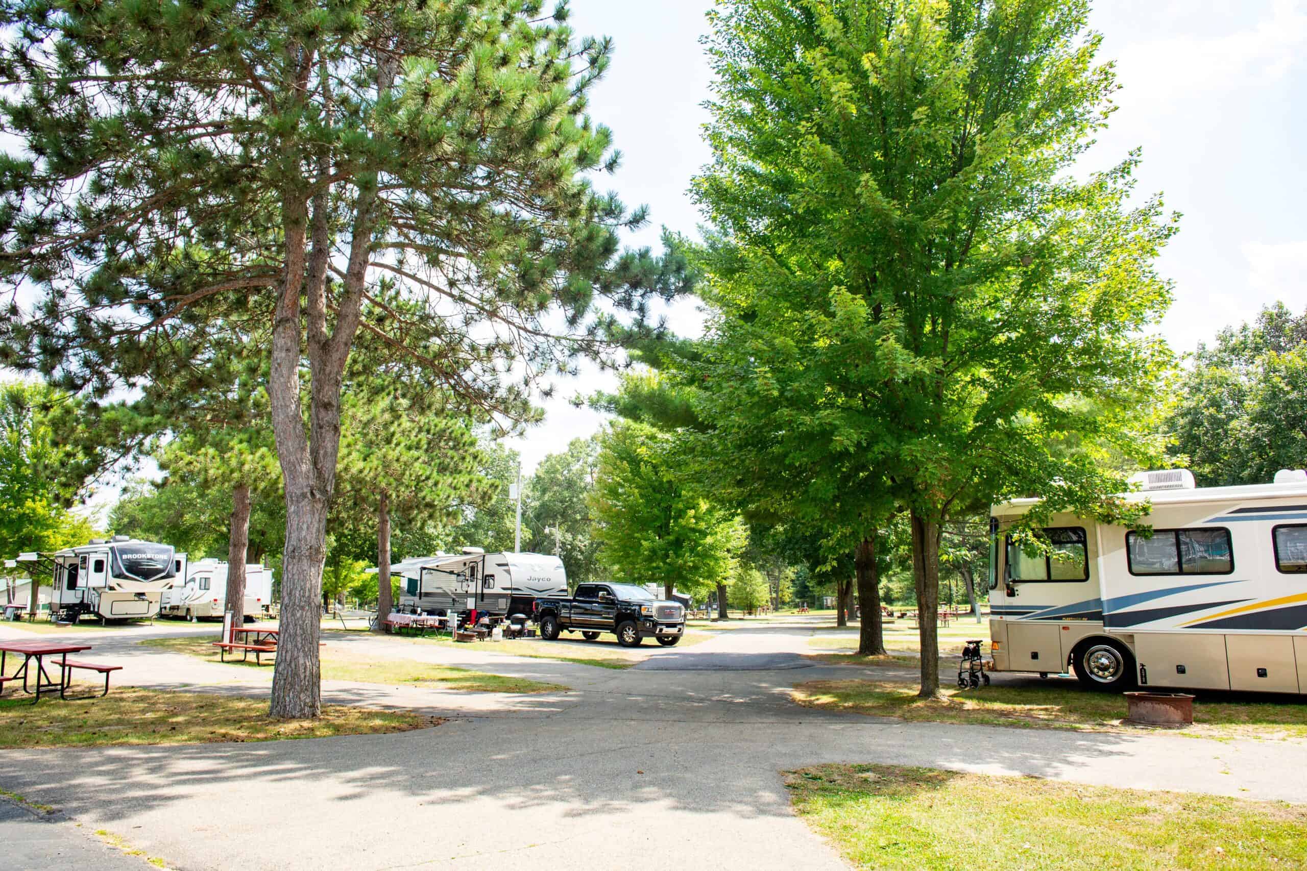 RV campground with several campers and trucks parked among tall trees, featuring empty picnic tables and paved roads under a clear sky.