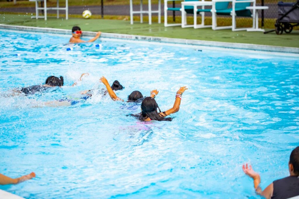 Several children are swimming in an outdoor pool, some with their arms outstretched, while another child watches from the poolside.