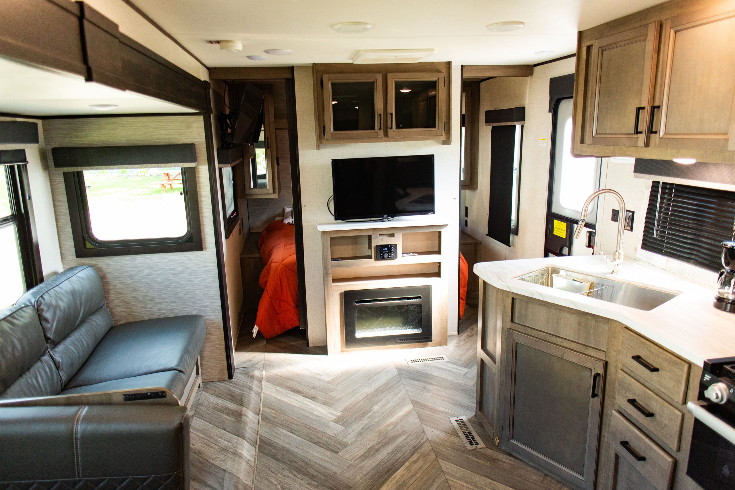 Interior of a modern RV with a small kitchen, sofa, wall-mounted TV, fireplace, and a bedroom with orange bedding visible in the background.