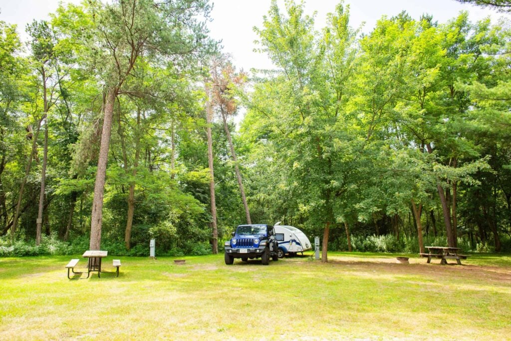 A blue SUV hitched to a white camper is parked on grass in a wooded campsite with picnic tables and trees.